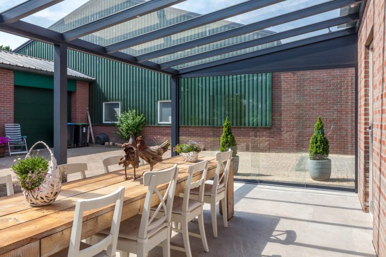 Terrassenüberdachung Pigato mit Glas Holzterrasse mit Esstisch, Stühlen und Pflanzen unter einem transparenten Dach.