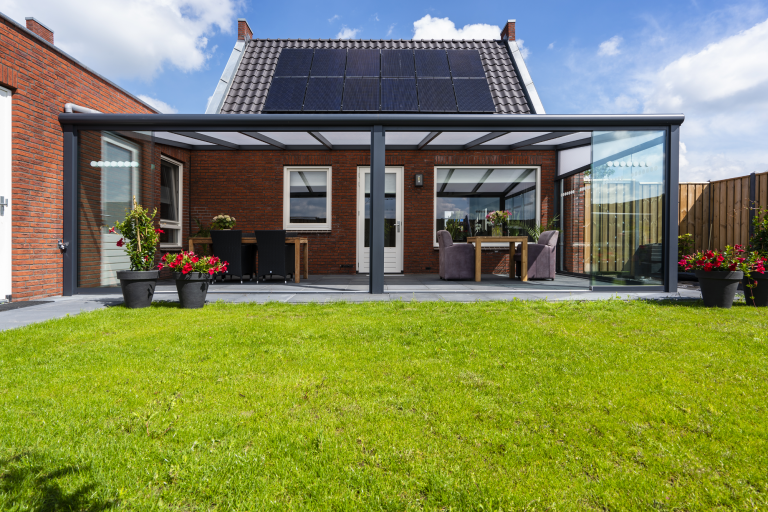 Terrassenüberdachung Bosco aus Aluminium mit Polycarbonat Modernes Haus mit Terrassenüberdachung und gepflegtem Rasen, umgeben von Pflanzen.